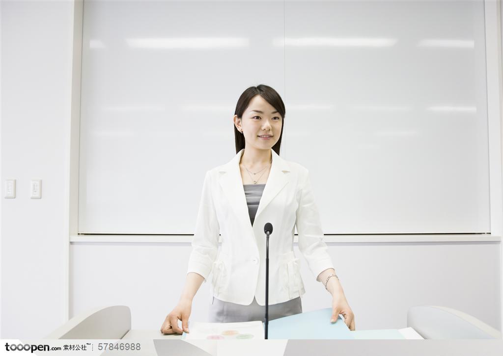 商业培训-穿白色西装在讲台话筒前演讲的美女老师