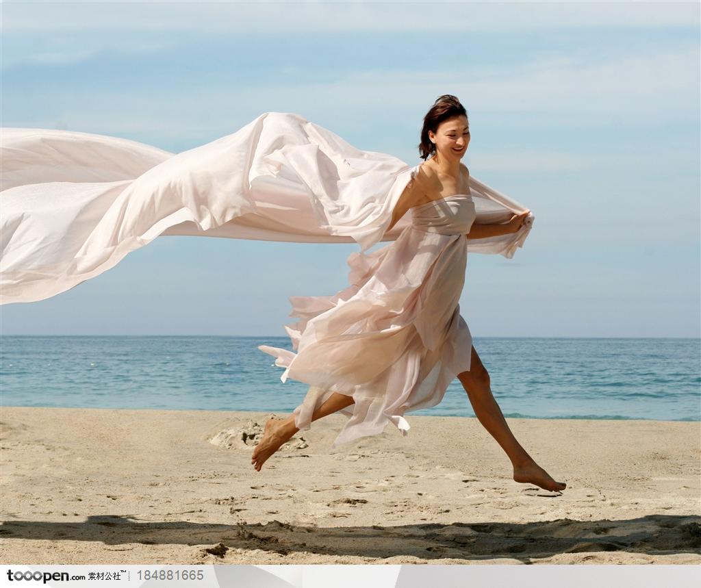 穿白色抹裙飘扬的披肩的美女双脚腾空在沙滩上跳跃